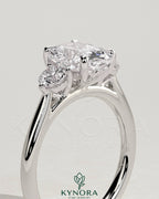 Kynora CJ elongated 2ct cushion claw trilogy engagement ring with round side stones and delicate lily detail in platinum setting.