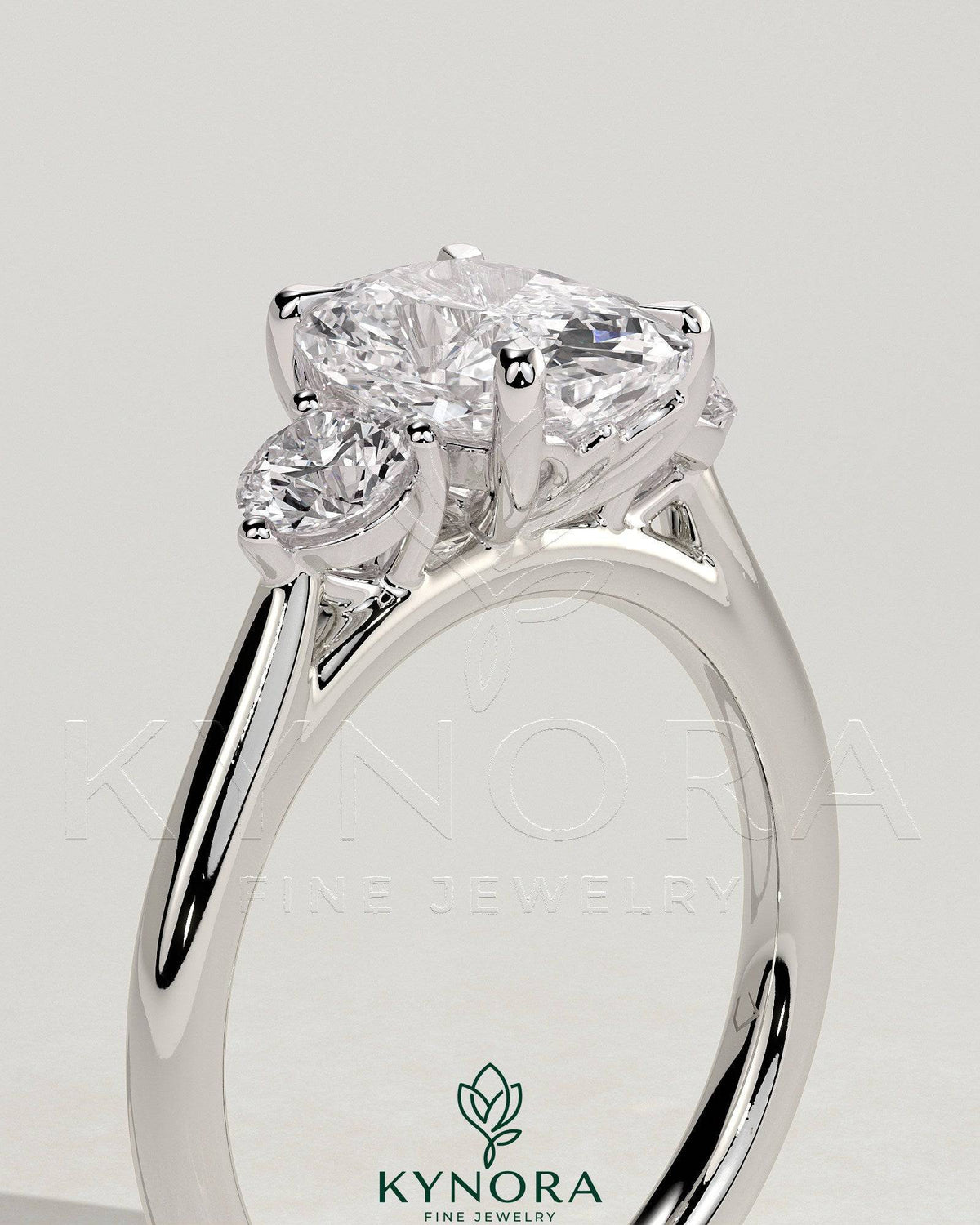 Kynora CJ elongated 2ct cushion claw trilogy engagement ring with round side stones and delicate lily detail in platinum setting.