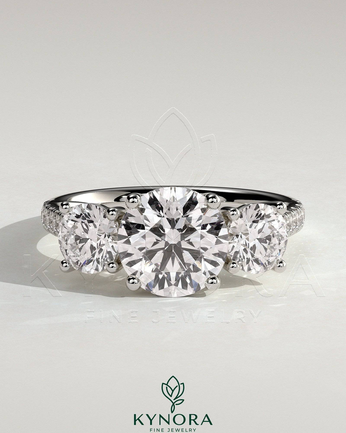 Kynora CJ three-stone engagement ring with 2ct round claws, side stones, pave band, and trellis gallery design.