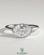 East-West 2-carat oval bezel solitaire engagement ring by Kynora CJ featuring a prominent oval diamond set horizontally on a sleek band