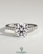Kynora CJ 2ct round solitaire engagement ring with hidden halo, pave band, and cathedral high setting, showcasing intricate jewelry design.