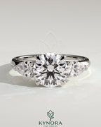 Kynora CJ three-stone engagement ring featuring a 2-carat round claw trilogy center with pear side stones and a knife edge band.