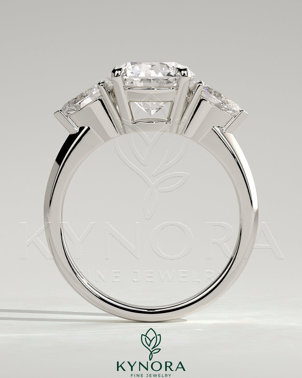 Kynora CJ 2-carat round claw trilogy engagement ring with pear side stones and knife edge band, featuring a sparkling three-stone design.