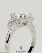 Kynora CJ 2ct round claw trilogy engagement ring with pear side stones and knife edge band, sparkling and elegant jewelry piece.